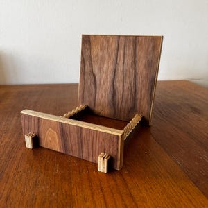 Puede incluir: Un soporte de exhibición de madera con una parte trasera rectangular y una base horizontal. El soporte está hecho de madera marrón oscuro con un patrón de vetas visible. La base tiene dos pequeños soportes cuadrados. El soporte está diseñado para sostener un objeto pequeño.