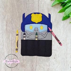 May include: A felt crayon holder shaped like a robot, featuring a blue helmet and a yellow badge. It has slots for crayons, with three crayons visible. A pink, yellow, and green crayon are placed near the holder. The holder is on a wooden surface.