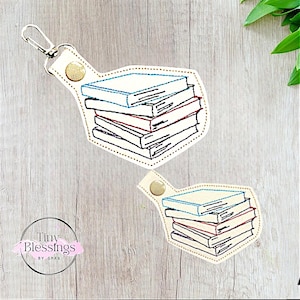 Peut inclure: Un porte-clés en cuir blanc avec une pile de livres brodés dessus. Le porte-clés a un anneau en métal doré et un fermoir en métal doré. Le texte "Tiny Blessings by G.M.A.B." est imprimé sur le porte-clés.