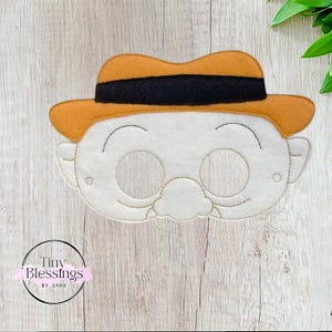 Puede incluir: Máscara de fieltro de un personaje de dibujos animados con bigote y sombrero. La máscara tiene agujeros para los ojos y es beige con detalles marrones y negros.