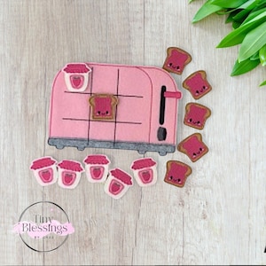 Puede incluir: Un juego de tic-tac-toe de fieltro rosa con un diseño de tostadora, con rebanadas de tostadas con mermelada y pequeños tarros de mermelada. El juego incluye el texto "Tiny Blessings by GMB".