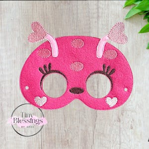 Op de afbeelding: Roze vilt lieveheersbeestje masker met zwarte ogen, bruine neus en roze hart accenten. Het masker heeft twee roze vilt antennes met hartvormen aan de uiteinden.