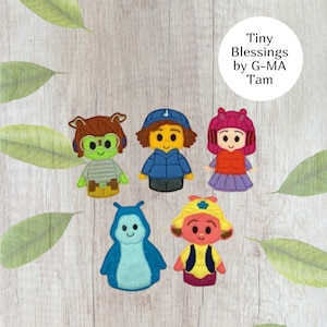 May include: Five colorful felt character dolls with various outfits and accessories, including hats and jackets. The dolls are arranged on a light wood-grain surface with green leaf accents. The text "Tiny Blessings by G-MA Tam" is in a white circle.