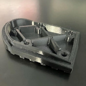 Peut inclure: Un objet noir imprimé en 3D de forme singulière. Il présente un bord incurvé avec des rainures et plusieurs supports et trous internes. L'objet semble être en plastique et repose sur une surface sombre.