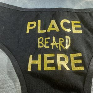 May include: Black underwear with gold lettering that reads "PLACE BEARD HERE". The text is centered on the front of the garment. The underwear has a thin waistband and trim.