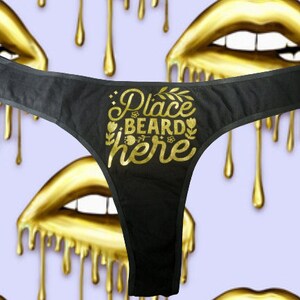 May include: Black thong underwear with gold text reading "Place Beard Here", surrounded by floral and leaf designs. The background has a repeating pattern of gold lips with dripping gold accents.