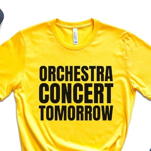 May include: A yellow t-shirt with the text "ORCHESTRA CONCERT TOMORROW" in bold black letters. The shirt is styled with black sunglasses, a black Adidas cap, black sneakers, a watch, jeans, and a small potted plant.