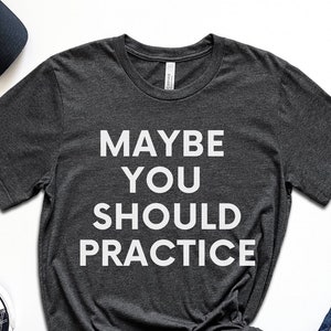 May include: Dark gray t-shirt with the text "MAYBE YOU SHOULD PRACTICE" in white. The image also includes black sunglasses, a black baseball cap, black sneakers, blue jeans, a succulent plant, a smartwatch, and white earbuds.