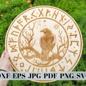 May include: A round, light-colored wooden plaque featuring a detailed raven perched on a tree branch. The design includes runes and celestial symbols. The text "DXF EPS JPG PDF PNG SVG" is at the bottom.