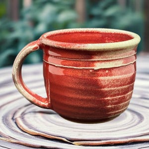Könnte beinhalten: Eine handgefertigte rote Keramik-Tasse mit Henkel. Die Tasse hat eine strukturierte Oberfläche mit horizontalen Linien und einen cremefarbenen Rand. Die Tasse steht auf einer dekorativen, kreisförmigen Oberfläche.