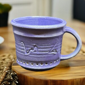 May include: A handcrafted lavender ceramic mug with a rounded handle. The mug features horizontal grooves and a decorative design. The base has small, evenly spaced holes. The mug sits on a wooden coaster.