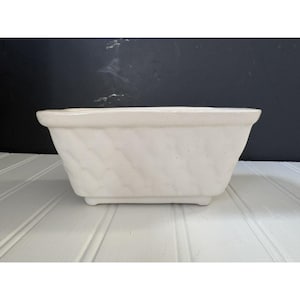 May include: Cream-colored rectangular planter with a textured, woven-like pattern on the sides. The planter has a slightly flared top and sits on four small feet. The background is a dark wall and a white-painted surface.