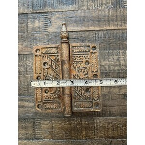Peut inclure: Une ancienne charni&egrave;re m&eacute;tallique orn&eacute;e avec un motif d&eacute;coratif. La charni&egrave;re est de couleur marron rouille et mesure environ 13 cm de long. La charni&egrave;re est ouverte, avec une r&egrave;gle pos&eacute;e dessus.
