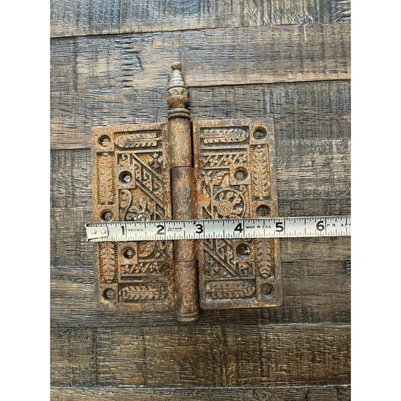 Peut inclure: Une ancienne charni&egrave;re m&eacute;tallique orn&eacute;e avec un motif d&eacute;coratif. La charni&egrave;re est de couleur marron rouille et mesure environ 13 cm de long. La charni&egrave;re est ouverte, avec une r&egrave;gle pos&eacute;e dessus.