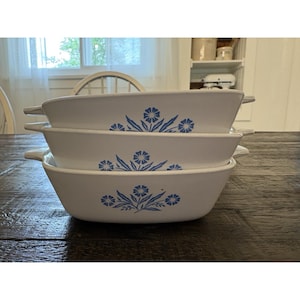 May include: Three white rectangular casserole dishes stacked on a dark wooden table. Each dish features a blue floral design. The dishes have handles on each side. The background includes a window and a white shelf.