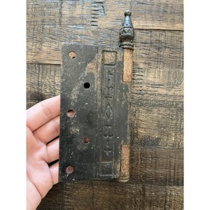 Peut inclure: Une ancienne charni&egrave;re en m&eacute;tal noir avec des d&eacute;tails d&eacute;coratifs et un aspect rouill&eacute;. La charni&egrave;re comporte des trous de montage et un fleuron d&eacute;coratif. Probablement pour une porte ou une armoire. Mesure environ 15 cm de long.