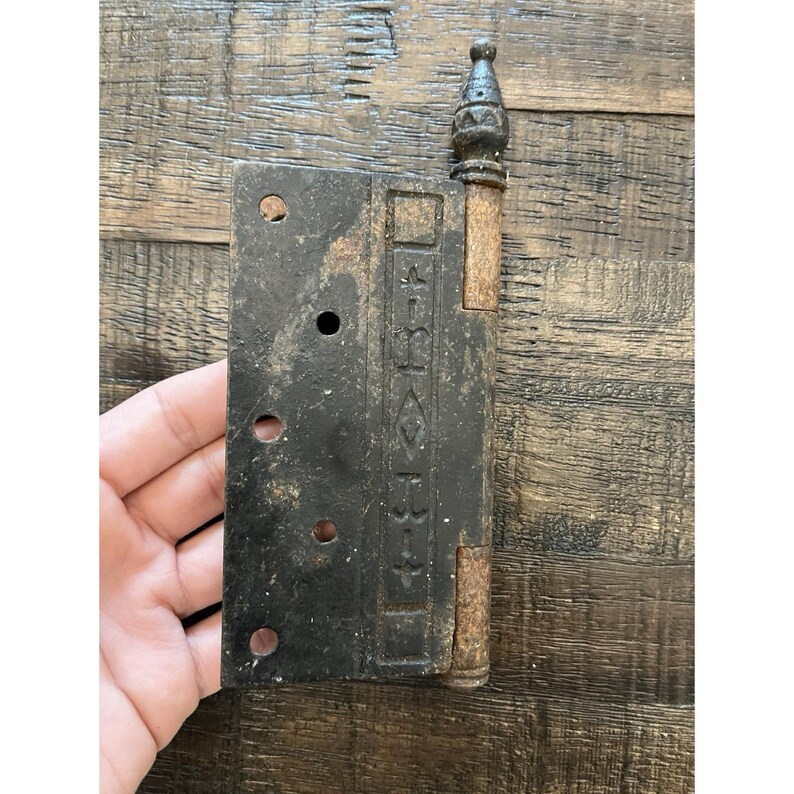 Peut inclure: Une ancienne charni&egrave;re en m&eacute;tal noir avec des d&eacute;tails d&eacute;coratifs et un aspect rouill&eacute;. La charni&egrave;re comporte des trous de montage et un fleuron d&eacute;coratif. Probablement pour une porte ou une armoire. Mesure environ 15 cm de long.