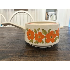 May include: A cream-colored ceramic saucepan featuring a speckled finish and a vibrant floral pattern of orange flowers with green leaves. The cookware has a single handle on one side, a small tab handle on the opposite side, and a thin red rim.