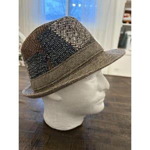 May include: A fedora hat with a patchwork design. The hat features panels of brown, black, and blue fabric with various textures, including herringbone and woven patterns. The brim and band are a lighter brown with a herringbone pattern.