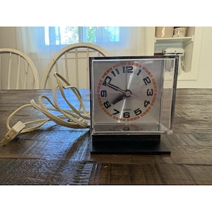 May include: A vintage square alarm clock with a silver face and orange numerals. The clock is encased in a clear plastic box and sits on a black base. A cream-colored cord with a plug extends from the clock.