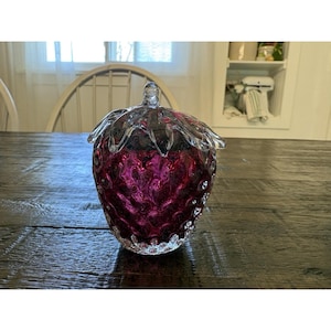 May include: A decorative glass jar shaped like a strawberry. The jar is a deep magenta color with a clear, textured lid. The jar sits on a dark wooden table.