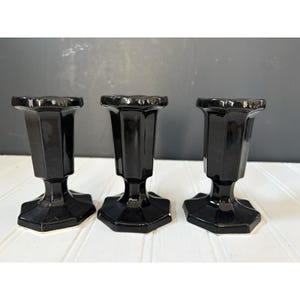 May include: Three black candle holders with a glossy finish. Each holder has a hexagonal base and a flared top with a scalloped edge. The holders are arranged in a row against a neutral background.