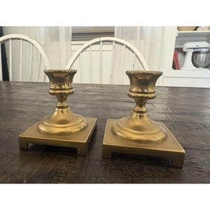 May include: Two antique brass candle holders. Each holder has a square base and a detailed, ornate design. The candle holders are a warm gold color and are displayed on a dark wooden surface.