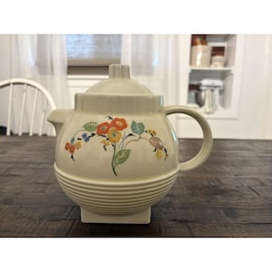 May include: A cream-colored ceramic teapot featuring a rounded body with horizontal ribbed detailing at the base and a matching lid. The front is decorated with a floral pattern in orange, yellow, blue, and green, and it has a curved handle.