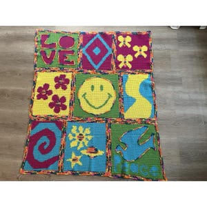 May include: A colorful, handmade crochet blanket featuring nine squares with various designs. The squares include the word "LOVE", a smiley face, flowers, a dove, and abstract shapes. The blanket is bordered with a contrasting color.