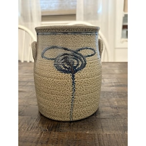 May include: A cylindrical stoneware crock with a textured tan surface and two side handles. The exterior features a hand-painted dark blue floral design consisting of a central spiral and three petals, with a vertical line extending downward.