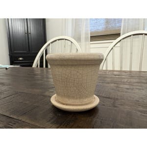 May include: A beige ceramic flower pot with a matching saucer. The pot has a cracked, textured surface. The pot is sitting on a dark wooden table.