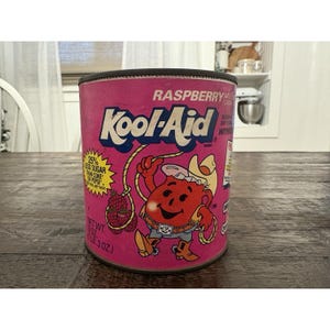 May include: A vintage raspberry Kool-Aid can with a pink and black design. The can features the Kool-Aid Man character, a lasso, and text that reads "25% less sugar than Coke or Pepsi." The can weighs 8.3 oz.
