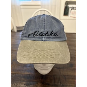 May include: A blue and tan baseball cap with the word "Alaska" embroidered in black script. The cap has a curved brim and a fabric strap for adjustment. The hat is made of a soft, worn material.