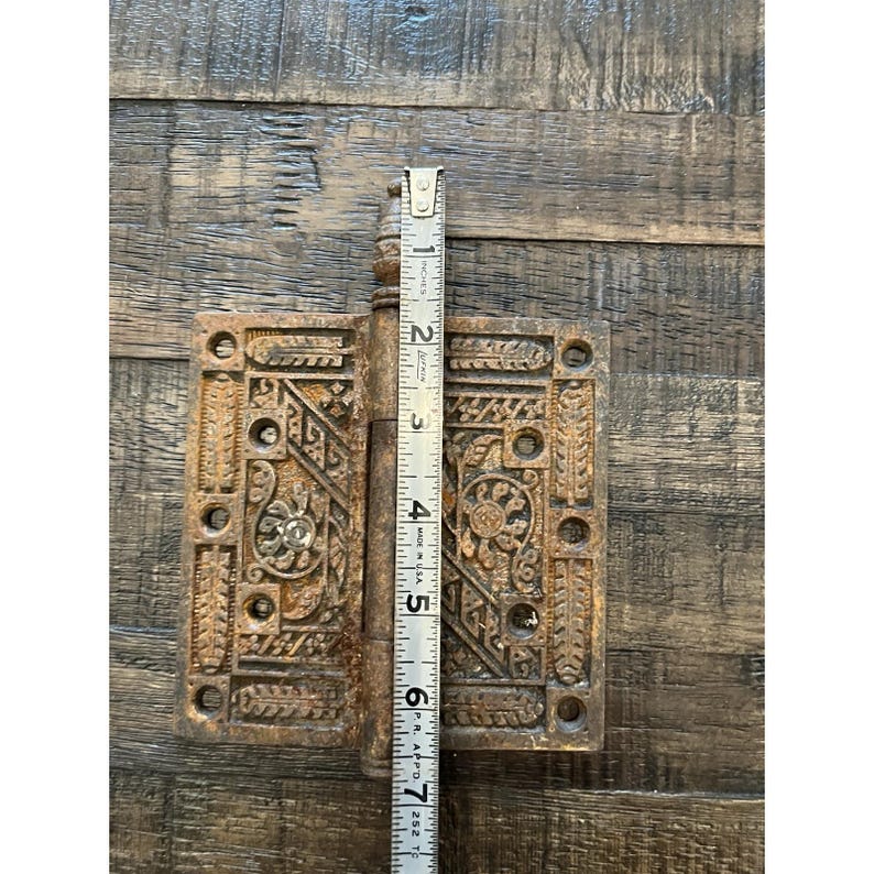 Peut inclure: Une ancienne charni&egrave;re de porte en bronze orn&eacute;e de motifs floraux et g&eacute;om&eacute;triques d&eacute;taill&eacute;s. La charni&egrave;re mesure environ 15 cm de hauteur, mesur&eacute;e par une r&egrave;gle. La charni&egrave;re est ouverte, r&eacute;v&eacute;lant son design et sa construction complexes.