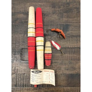 May include: Three cylindrical fishing floats with red, white, and yellow painted bands, alongside two metal fishing lures with hooks. A paper instruction card labeled Dayton Fishing Floats is included, detailing usage for bait depth regulation.
