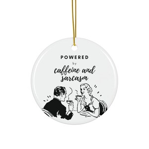 Op de afbeelding: Witte keramische ornament met een zwart-witte illustratie van een stel dat koffie drinkt. De tekst "POWERED by caffeine and sarcasm" is op de ornament gedrukt.