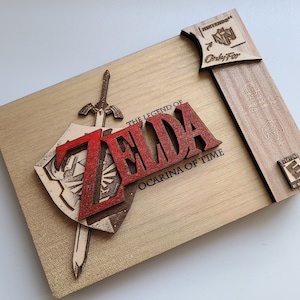 May include: A wooden box featuring a 3D design of the Legend of Zelda: Ocarina of Time logo. The logo is red and includes a sword and shield. The box is made from light brown wood and has a Nintendo 64 logo on the side.