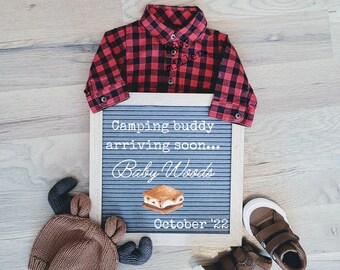 Può includere: Una camicia da bambino a quadri rossi e neri, un cappello di lana marrone con le corna, un paio di scarpe da bambino in pelle marrone e una lavagna con le lettere con il testo "Camping buddy arriving soon... Baby Woods October '22" e un disegno di un s'more.