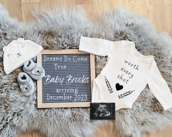 May include: An announcement of a baby's arrival, featuring a white onesie with "worth every shot" text, a letter board with "Dreams Do Come True... Baby Brooks arriving December 2023", a baby hat, booties, and an ultrasound image.