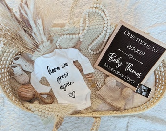 Op de afbeelding: Een baby aankondigingsarrangement in een gevlochten mand. Het bevat een witte romper met de tekst "here we grow again", een letterbord met "One more to adore! Baby Thomas November 2023", een gebreide muts, babyschoentjes en een echo.