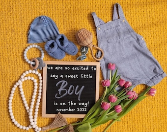 Może przedstawiać: Tablica z napisem "we are so excited to say a sweet little Boy is on the way! november 2022". Tablica jest otoczona przedmiotami dla dzieci, w tym niebieską czapką w białe paski, niebieskimi bucikami dla niemowląt, drewnianym grzechotką, białym naszyjnikiem z koralików i różowymi tulipanami.