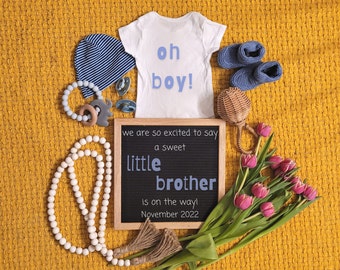 Może przedstawiać: Biały śpioszek z napisem "oh boy!" w niebieskich literach, tablica z literami z napisem "we are so excited to say a sweet little brother is on the way! November 2022", niebiesko-biała czapka w paski, niebieska grzechotka, drewniana grzechotka, bukiet różowych kwiatów i dwa niebieskie buciki dla niemowlęcia.
