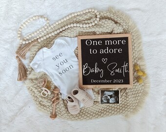 Op de afbeelding: Een witte romper met de tekst "see you soon" erop, een letterbord met de tekst "One more to adore Baby Smith December 2023", een houten rammelaar, een paar gehaakte babyschoentjes, een houten kralensnoer en een echo-foto op een beige pluche deken.