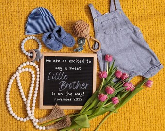 Puede incluir: Un tablero de letras negro con el texto "we are so excited to say a sweet Little Brother is on the way! november 2022". El tablero de letras está rodeado de una variedad de artículos para bebés, que incluyen un gorro a rayas azul y blanco, un par de zapatos de bebé azules, un sonajero de madera, un collar de cuentas blancas y un ramo de flores rosas.