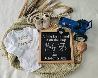 May include: A baby announcement featuring a white onesie with "Tiny Farmer" in gold lettering, a chalkboard sign, and a blue toy tractor. The sign reads "A little farm hand is on the way Baby Ellis due October 2022."