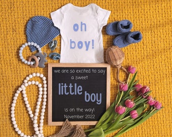 Może przedstawiać: Biały śpioszek z napisem "oh boy!" w niebieskich literach, niebiesko-biała czapka w paski, tablica z literami z napisem "we are so excited to say a sweet little boy is on the way! November 2022", drewniana grzechotka, sznur białych koralików i bukiet różowych tulipanów.