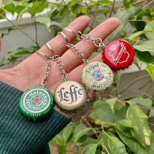 Peut inclure: Quatre porte-clés en forme de capsule de bouteille avec différents logos de marques. Les porte-clés sont vert, or, beige et rouge. Les logos sont Heineken, Leffe, Tripel Karmeliet et Smirnoff.