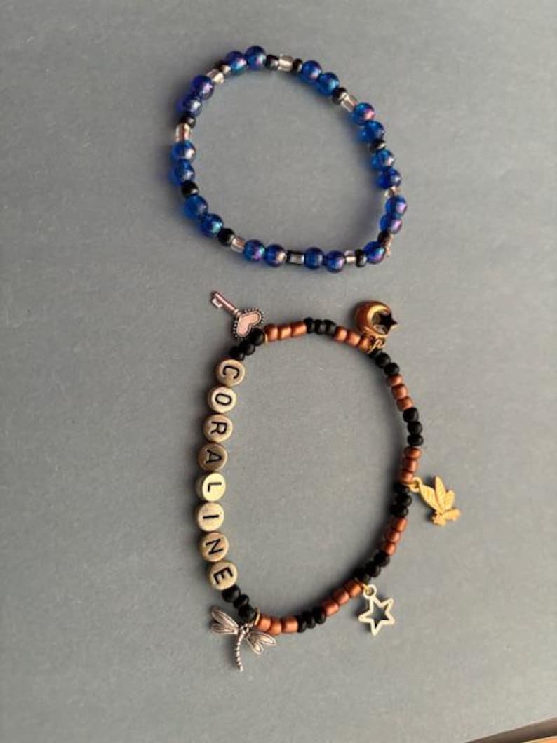 May include: Two bracelets, one blue with silver beads and one brown with black beads and silver charms. The brown bracelet has the name "CORALINE" spelled out in gold beads.