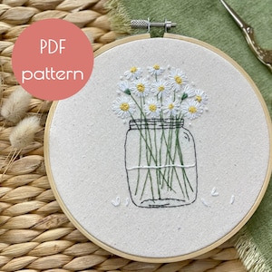 May include: A white embroidery hoop with a hand-stitched design of a glass jar filled with a bouquet of white daisies with yellow centers. The embroidery is on a white fabric background.