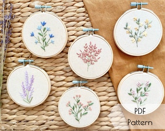 May include: Five embroidery hoop patterns featuring different floral designs. The hoops are arranged in a semi-circle on a woven straw surface. The designs include blue flowers, pink flowers, yellow flowers, lavender, and white flowers. The text "PDF Pattern" is visible at the bottom of the image.
